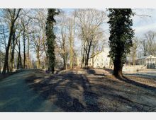 Weitläufiger Park mit hohen, teils efeubewachsenen Bäumen und kahlen Ästen im Winter. Im Hintergrund ist ein historisches, helles Gebäude mit mehreren Fenstern zu sehen, daneben kleinere Gebäude und ein Wasserlauf.