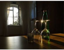 Ein heller Raum mit einem großen Fenster, durch das Sonnenlicht einfällt, und weißen Vorhängen. Im Vordergrund stehen eine grüne Wasserflasche, zwei umgedrehte Weingläser und ein kleines Glas auf einem Holztisch.