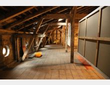 Ein Dachboden mit sichtbaren Holzbalken und einem Holzboden, beleuchtet durch kleine runde Fenster in den Wänden. Im Raum befinden sich vereinzelte Gegenstände wie eine orangefarbene Schüssel und Trennwände aus Holzrahmen und Platten.