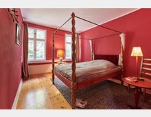 Schlafzimmer mit roten Wänden, einem Himmelbett aus Holz und einem Holzfußboden. Zwei große Fenster lassen natürliches Licht herein, während eine Nachttischlampe und ein Beistelltisch mit Büchern und dekorativen Elementen das Zimmer ergänzen.