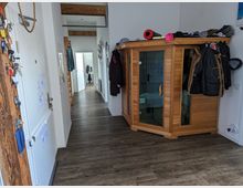 Flur einer Wohnung mit Holzboden, einer Innenkabine aus Holz (vermutlich eine Sauna) an der rechten Wand und mehreren aufgehängten Jacken. Links befinden sich eine Tür und ein Schlüsselbrett, im Hintergrund führt der Flur zu weiteren Räumen mit weiß gestrichenen Wänden und Sichtbalken.