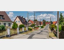 Eine ruhige Wohnstraße mit Einfamilienhäusern, die teils verputzt oder mit Klinker verkleidet sind und rote, braune oder graue Dächer haben. Die Häuser stehen in Gärten mit Zäunen, und entlang der Straße befinden sich Strommasten sowie vereinzelte Bäume.