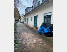 Schmaler, gepflasterter Hofweg entlang eines weißen Wohnhauses mit grün gerahmten Fenstern und grüner Haustür sowie Satteldach. Am Rand stehen Pflanzen und ein abgedecktes Fahrzeug; der Weg führt zu weiteren Gebäuden und Bäumen im Hintergrund.