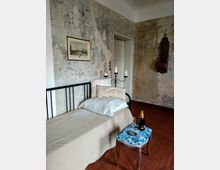 Ein rustikales Zimmer mit einer einzelnen Metallbettgestell, das mit beigen Kissen und einer Decke ausgestattet ist. Die Wände sind verputzt und zeigen Abnutzungsspuren, ein gerahmtes Bild hängt an der Wand, und darunter steht ein kleiner Hocker mit blauer Polsterung, auf dem eine Flasche Champagner und ein Glas stehen.