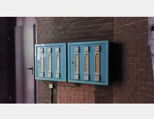 Zwei blaue, rechteckige Sicherungskästen mit vertikalen Schaltern, montiert an einer Wand aus roten Ziegelsteinen. Links daneben befindet sich eine geschlossene Tür mit einem schwarzen Türgriff.