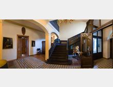 Eingangsbereich eines historischen Gebäudes mit einer hölzernen, dunklen Treppe, dekorativen Fliesen am Boden und Glastüren auf der rechten Seite. An den Wänden hängen Portraitgemälde, und neben der Treppe steht ein Holzspielzeugpferd.