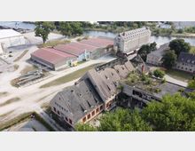 Luftaufnahme eines Industriegeländes mit verfallenen Gebäuden, darunter ein großes Haus mit teilweise eingestürztem Dach und Bewuchs. Im Hintergrund sind Lagerhallen mit roten Dächern, ein Fluss und weitere Wirtschaftsgebäude zu sehen.