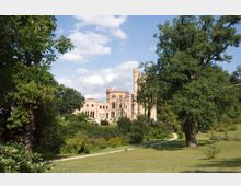 Historisches Schloss mit hellen Steinmauern und einem hohen Rundturm, umgeben von großen Bäumen und einer weitläufigen, gepflegten Parklandschaft. Ein schmaler Weg führt durch die Wiese im Vordergrund.