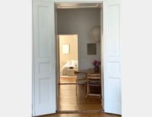 Blick durch geöffnete weiße Flügeltüren in einen Wohnraum mit Holzboden; im Vordergrund ein runder Holztisch mit Stühlen und einer Blumenvase, im Hintergrund ein Schlafzimmer mit Bett und heller Bettwäsche.