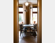 Ein gemütlicher Besprechungs- oder Essbereich mit einem langen Tisch und einer Mischung aus verschiedenen Stühlen in einem Raum mit holzfarbenem Boden. Große Fenster mit weißen Vorhängen lassen Tageslicht einfallen, während hängende, kugelförmige Lampen den Raum beleuchten.