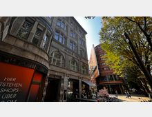 Straßenszene mit dem historischen Gebäude "Haus Huth" aus Stein mit Rundbogenfenstern links und einem modernen, rotbraunen Gebäude mit gläsernem Vordach rechts. Dazwischen eine kleine Außengastronomie und im Hintergrund Bäume in einer Fußgängerzone.