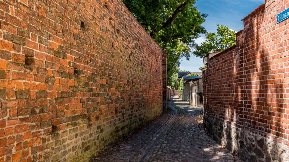 Schmale Kopfsteinpflastergasse zwischen hohen roten Backsteinmauern, die in Richtung kleiner Gebäude und eines Eingangstores führt. Oben rechts ist ein Straßenschild „Glintzmauer“ zu sehen, darüber ragen Bäume in den Himmel.