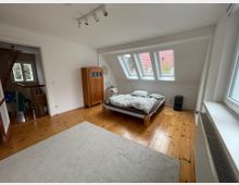 Helles Dachgeschoss-Schlafzimmer mit Holzfußboden, niedrigem Bett und Kleiderschrank. An der schrägen Dachwand befinden sich mehrere Dachfenster, daneben ein großes Seitenfenster und ein offener Durchgang zu einem angrenzenden Raum.