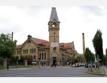 Backsteingebäude im Stil der Industriearchitektur mit spitzem Turm, erkennbar als "Kulturbrauerei". Im Vordergrund eine Kreuzung, Bäume und geparkte Fahrzeuge, im Hintergrund ein Schornstein.