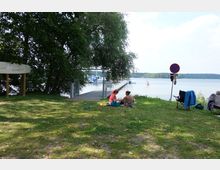 Grüne Wiese mit Bäumen am Ufer eines Sees, im Hintergrund ein Steg mit Booten und eine schwimmende Boje. Im Vordergrund sitzen Personen auf der Wiese oder in Campingstühlen, ein Verkehrsschild steht am rechten Bildrand.