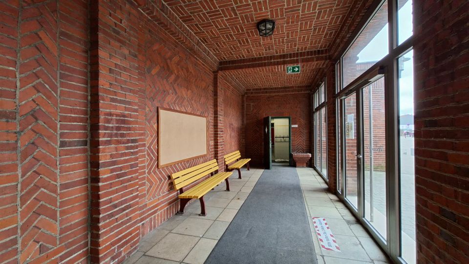 Überdachter Eingangsflur aus rotem Backstein mit gemustertem Ziegelgewölbe, zwei gelben Holzbänken und grauem Teppichläufer. Rechts verläuft eine Glasfront mit Türen nach außen, am Ende führt eine Tür in den Innenbereich.