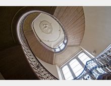 Ansicht einer spiralförmigen Treppe von unten, mit kunstvoll verziertem Geländer und einem ovalen Deckenrelief. Der Raum wird durch große Fenster mit Tageslicht erhellt.