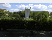 Ein Park mit einer Bank im Vordergrund, umgeben von Bäumen und Sträuchern. Im Hintergrund steht ein großes Betondenkmal mit nach oben ragenden Elementen, umgeben von einer Grünfläche und weiteren Bäumen.