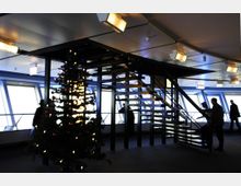 Innenraum mit moderner Architektur, einer offenen Metalltreppe in der Mitte und großen Fenstern, die diffuses Licht hereinlassen. Ein geschmückter Weihnachtsbaum steht im Vordergrund, und mehrere Personen bewegen sich durch den Raum.