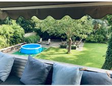 Ein gepflegter Garten mit einer Rasenfläche, einem aufblasbaren blauen Pool, einer Sitzfläche mit Holzstühlen auf einer Holzterrasse unter einem Baum sowie dichten, grünen Pflanzen im Hintergrund. Im Vordergrund ist ein überdachter Bereich mit Sitzkissen zu sehen.