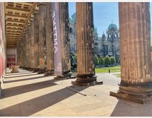 Kolonnade mit massiven Steinsäulen des Alten Museums in Berlin, mit einem Banner mit der Aufschrift „Antike“ und Blick auf den Rasenplatz und die Kuppel des Berliner Doms im Hintergrund bei sonnigem Wetter.
