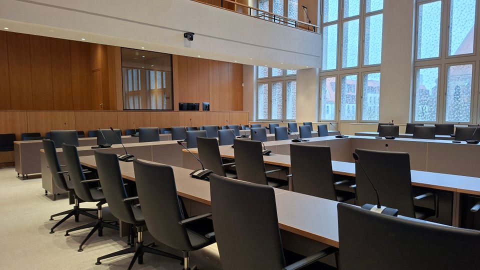 Moderner Sitzungssaal mit U-förmig angeordneten Tischen, schwarzen Drehstühlen und integrierten Mikrofonen. Hohe Fensterfronten und Holzvertäfelung an den Wänden prägen den hellen Raum.