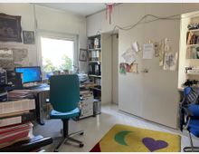 Ein kleines Büro mit einem Schreibtisch, Computer, Monitor und einem grünen Drehstuhl in der Mitte. An den Wänden befinden sich Regale mit Ordnern und Dokumenten, sowie ein großer Schrank mit angepinnten Zetteln. Ein Fenster lässt Tageslicht herein, darunter steht eine Pflanze. Der Boden ist mit einem gelben Teppich mit buntem Mond- und Herzmotiv bedeckt.