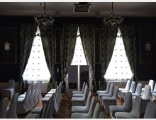 Raum mit Reihen weiß überzogener Stühle, großen Fenstern mit dekorativen, dunkelgrünen Vorhängen und zwei hängenden Kronleuchtern. Die Wände sind dunkel gestrichen und mit goldgerahmten Gemälden verziert.