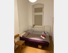 Ein einfacher Schlafraum mit einem Doppelbett, darauf graue Bettwäsche und gefaltete Handtücher in verschiedenen Farben (weiß, blau, rot). Im Raum befinden sich ein Holzstuhl, ein Heizkörper unter einem Fenster mit heruntergelassenem Rollo, sowie eine kleine Leselampe auf einem Regal am Kopfende des Bettes.