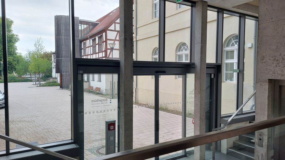 Blick aus einem modernen Gebäudeeingang mit breiten Glastüren und Betonpfeilern auf einen Innenhof. Im Hintergrund sind ein Fachwerkgebäude sowie ein gelbes Gebäude mit Rundbogenfenstern zu sehen, umgeben von Pflasterflächen und Grünbereichen.