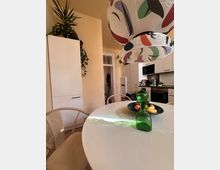 Helle moderne Küche mit weißer Einrichtung, Holzmöbeln und einem runden Esstisch mit einer grünen Glaskaraffe und einem Obstteller. Hängelampe mit buntem Muster und Pflanzen als Dekoration.