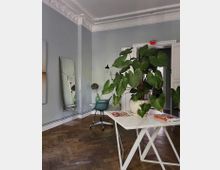 Heller Innenraum eines Zimmers mit hohen Decken und dekorativen Stuckverzierungen. Im Vordergrund steht ein weißer Tisch mit einer großen Zimmerpflanze, umgeben von Holzboden, einem grünen Schreibtischstuhl und einem großen Spiegel an einer hellgrauen Wand.