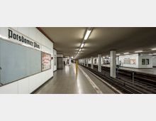 U-Bahn-Station mit dem Schriftzug „Potsdamer Platz“ an der weißen gefliesten Wand. Der Bahnhof hat zwei Gleise, mehrere Pfeiler und helle Deckenleuchten, mit Treppen und Rolltreppen im Hintergrund.