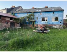 Hinterhof mit hohem Gras vor einem langgestreckten, blau verputzten Wohnhaus mit dunkel gedecktem Satteldach und mehreren Dachfenstern. Links steht ein niedriger Backstein-Nebengebäude mit Holztor, in der Mitte führt eine grüne Haustür ins Gebäude, daneben liegen eine Wiese und ein Picknicktisch.