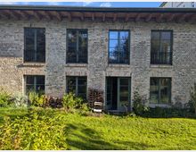 Backsteinfassade eines zweistöckigen Gebäudes mit großen, dunkel gerahmten Fenstern und einer Glastür im Erdgeschoss. Der Vordergrund besteht aus einem Garten mit Rasen, Sträuchern und gestapeltem Holz vor der Wand.