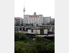 Ein Wasserkanal mit festgemachten Hausbooten im Vordergrund, umgeben von Gras und Büschen. Im Hintergrund sind ein historisches Gebäude mit rotem Dach sowie der Berliner Fernsehturm sichtbar.
