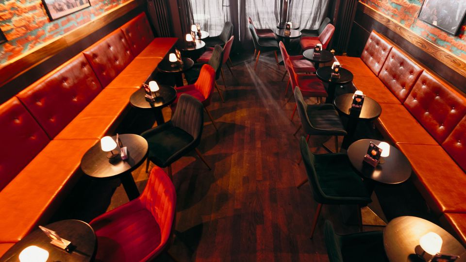 Schmaler Bar- oder Loungebereich mit langen, gepolsterten Lederbänken entlang beider Seiten, kleinen runden schwarzen Tischen mit Tischlampen und gemischten Polsterstühlen. Freiliegende Backsteinwände mit Bildern, dunkler Holzboden und bodenlange Vorhänge am Ende des Raums.