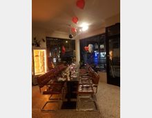 Ein modernes Restaurant mit einer langen Tafel, die für ein Abendessen gedeckt ist. Ledersessel mit Metallrahmen umgeben den Tisch, rote Herz-Ballons hängen von der Decke, und eine Glasvitrine mit beleuchteten Kuchen befindet sich links im Bild. Durch die Fenster ist die nächtliche Straßenbeleuchtung zu sehen.