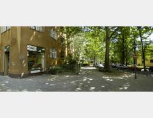 Straßenszene mit einer gelben Ecke eines Wohngebäudes im Vordergrund, das große Fenster mit Pflanzen zeigt und von Bäumen umgeben ist. Im Hintergrund befinden sich geparkte Autos entlang einer von Bäumen gesäumten Straße mit einem gelben Gebäude auf der gegenüberliegenden Seite.