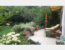 Ein gepflegter Garten mit einer gefliesten Terrasse, gesäumt von blühenden Blumen in Töpfen und Beeten. Im Hintergrund sind Sträucher, Bäume und ein oranger Sonnenschirm sowie eine Liege mit gelber Auflage zu sehen.