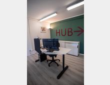Ein moderner Arbeitsbereich mit einem Schreibtisch, Bürostuhl und Monitor vor einer grünen Wand mit der Aufschrift "HUB" und einem Pfeil darauf. Der Raum hat einen hellen Laminatboden, eine weiße Wand mit einem Bild und moderne Deckenbeleuchtung.