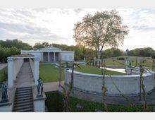 Klassizistisches Gebäude mit Säulenportikus inmitten eines Parks, umgeben von Rasenflächen und Symmetrie-elementen wie einer Pergola, Statuen und einem Wasserbecken. Im Hintergrund sind Bäume und offene Landschaft sichtbar.