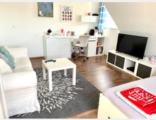 Helles Zimmer mit Holzboden, ausgestattet mit einem weißen Sofa, einem kleinen Couchtisch und einem grauen Teppich. Im Hintergrund befinden sich ein Schreibtisch mit Stuhl, Regale mit Dekoration sowie ein Fernseher auf einem weißen Medienmöbelstück vor einer Dachschräge.