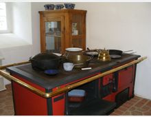 Ein rustikaler Raum mit einem alten roten Holzofen, der mit Küchengeräten wie Töpfen, Pfannen und einer messingfarbenen Teekanne dekoriert ist. Im Hintergrund steht ein Holzschrank mit Glasfront, in dem Geschirr aufbewahrt wird.