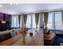 Wohnzimmer mit modernem Design, großer Holztisch in der Mitte, umgeben von gepolsterten Stühlen. Große Fenster mit hellen Vorhängen lassen viel Tageslicht herein; im Hintergrund Pflanzen und eine lilafarbene Wand mit einer Stehlampe.