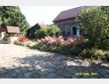 Hof mit gepflastertem Weg, einer Backsteinfassade mit roten Ziegeldach und einem Garten voller blühender Sträucher, darunter rosa Blumen; im Vordergrund ein Brunnen mit einem kleinen Dach.