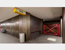 Eine U-Bahn-Station mit braunen Wandverkleidungen, einem gelben Schild mit der Aufschrift „Kiosk“ und einem roten Absperrgitter im Hintergrund. Links befinden sich Sitzbänke und ein Abfallbehälter.