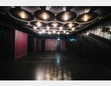 Ein moderner Innenraum mit dunklem Holzfußboden, dunkelvioletten Wänden und einer Decke mit dekorativen, runden Leuchtelementen. Die rechte Wand ist mit glänzenden schwarzen Kacheln verkleidet, und eine Tür befindet sich an der Rückwand.