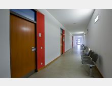 Langer, heller Flur in einem modernen Gebäude mit weißen Wänden und roten Akzentstreifen. Entlang einer Wand stehen mehrere graue Sitzreihen, während auf der gegenüberliegenden Seite Holztüren zu verschiedenen Räumen führen.