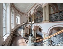 Großzügiges historisches Treppenhaus mit geschwungener Treppe, verzierten Geländern aus schwarzen Schmiedeeisen, Säulen und hohen Rundbogenfenstern. Die Decke ist mit Stuckornamenten geschmückt, und warme Pastellfarben dominieren den Raum.